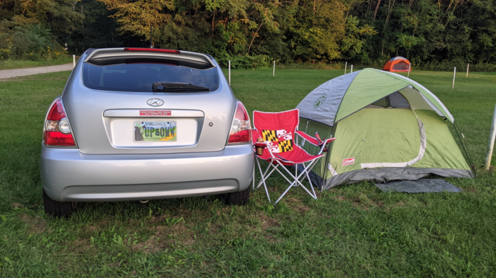 The back of a closed silver hatchback and a green tent with a Maryland state flag red, yellow, black, and white outdoor chair sitting in a field with a wooded area beyond.