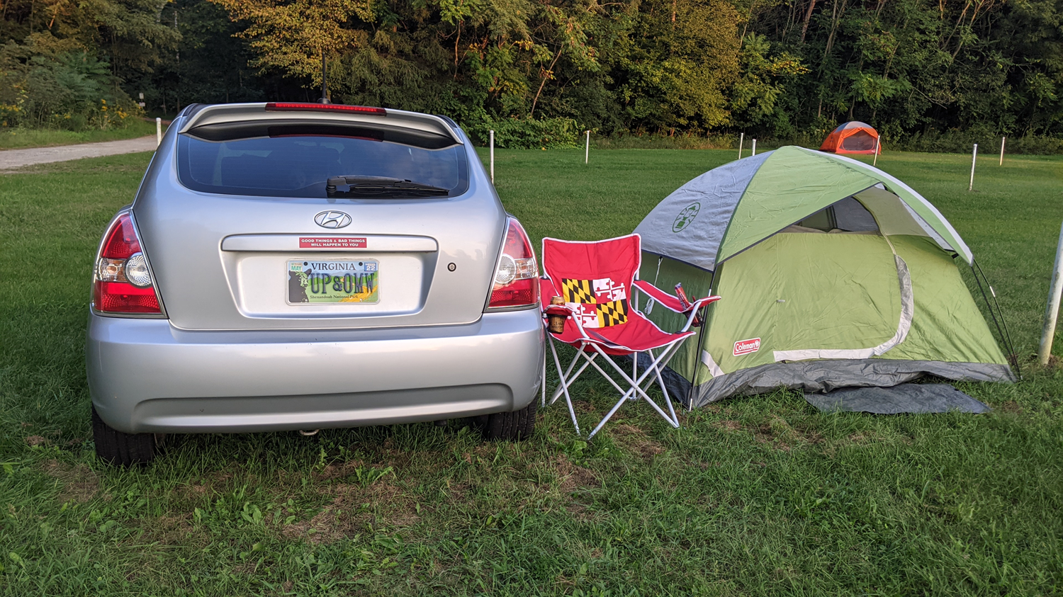 The back of a closed silver hatchback and a green tent with a Maryland state flag red, yellow, black, and white outdoor chair sitting in a field with a wooded area beyond.