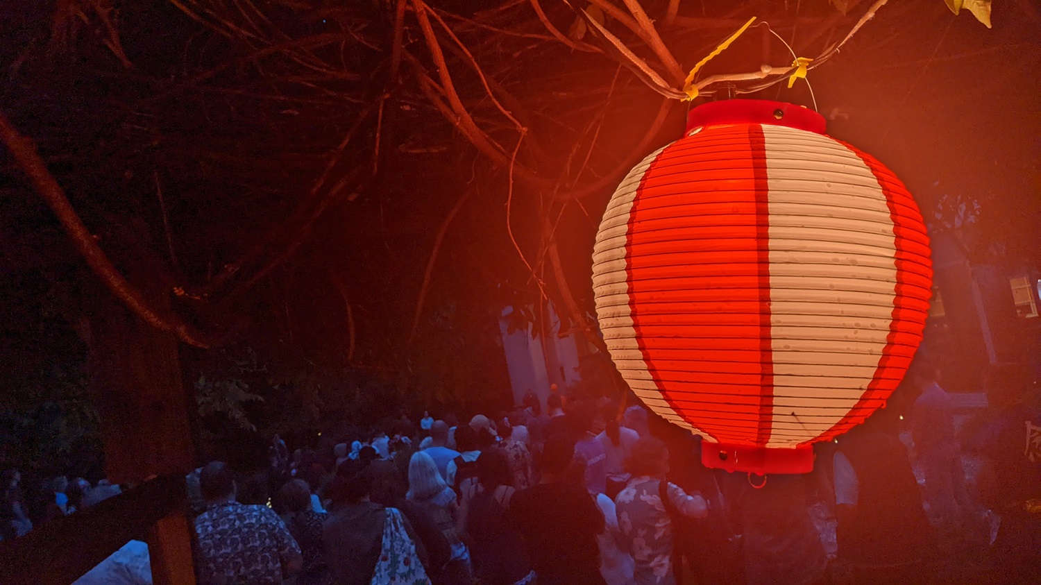 A red and white lamp glows in the foreground with people gathered for the summer Obon festival beyond in the darkness.